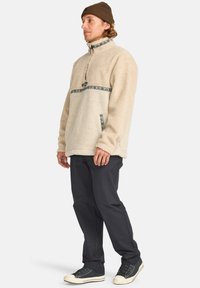 Beige fleece trøje med høj krave, lynlås i halv længde foran og lomme på forsiden. Blå mønstrede detaljer tværs over brystet. Sorte bukser og sko.
