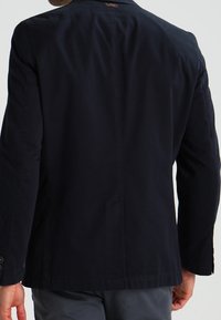 Granatowa bawełniana marynarka o dopasowanym kroju, z pojedynczym rozcięciem, mankietami na dwa guziki i gładką fakturą. Posiada minimalistyczne akcenty wzornicze.