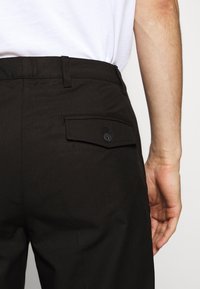 Černé kalhoty s kapsou na zadní straně, která má zapínání na knoflík. Vyrobeny z strukturované tkaniny, pas je plochý s poutky na opasek.