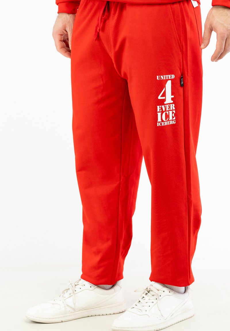 Røde træningsbukser lavet af glat stof, med et grafisk print med hvid tekst på venstre side, parret med hvide sneakers.