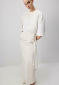 Top bianco a maniche corte abbinato a una gonna color crema lunga fino al pavimento, caratterizzata da un design avvolto e dettagli con bottoni dorati lungo la vita.