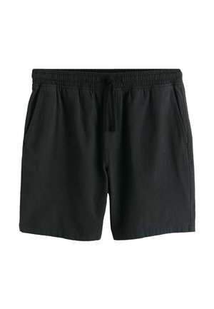 Férfi áll a fűben fekete rövidnadrágban, fehér zokniban, fekete-fehér sportcipőben és fehér pólóban, rajta színes grafikával.