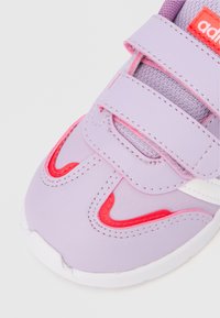 Λιλά αθλητικό παπούτσι με ροζ λεπτομέρειες και λευκά διακοσμητικά. Λουράκια με βέλκρο στο πάνω μέρος και υφασμάτινη επιφάνεια με υφή για άνεση. Στρογγυλεμένη μύτη και μαλακός δακτύλιος στον αστράγαλο.