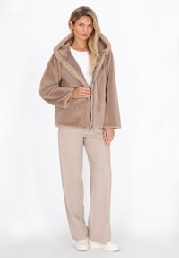 Giacca beige morbida con zip, texture pelosa e cappuccio, abbinata a pantaloni larghi beige chiaro e sneaker bianche.