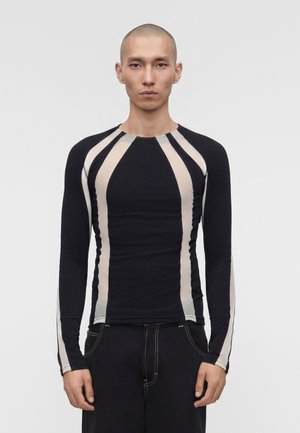 Homme au crâne rasé portant une chemise noire ajustée à manches longues avec des rayures verticales beige et un pantalon noir avec des coutures blanches, debout devant un fond blanc.