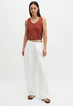 Vrouw staand, draagt een roestkleurig mouwloos topje, witte wijde broek met een beige riem en beige sandalen met strikjes.