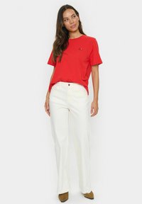 Woman wearing a red short-sleeve shirt with black text and high-waisted white pants, standing and looking to the side against a white background.