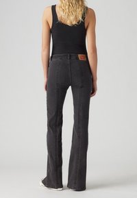 Jean en denim noir évasé avec des coutures visibles, une taille haute et un écusson en cuir avec logo à l'arrière. Porté avec un débardeur noir ajusté.