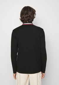 Svart långärmad polo-shirt med en kontrasterande krage som har röda, vita och svarta ränder. Slät tyg, avslappnad passform, rundad nederkant.