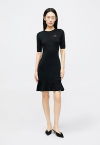 Vestido preto de malha com mangas curtas, textura canelada e barra com volantes. Apresenta um pequeno logótipo dourado no peito. Usado com saltos altos pretos.