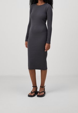 Szara sukienka z długim rękawem o dopasowanym kroju, wykonana z gładkiej tkaniny, sięgająca poniżej kolan, zestawiona z czarnymi sandałami na paski.