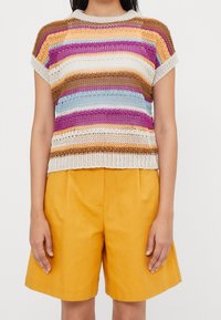 Top de ganchillo a rayas con líneas horizontales en marrón, amarillo, púrpura y azul, con un escote amplio y mangas cortas. Combinado con pantalones cortos mostaza.