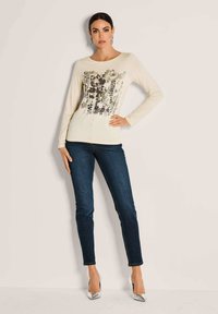 Cremfarbener Langarmtop mit abstraktem Muster in Grautönen und Braun, kombiniert mit dunkelblauen Skinny-Jeans und silbernen High Heels.