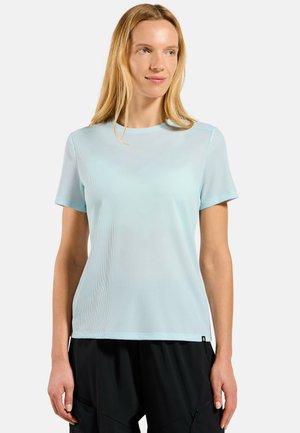 Femme aux longs cheveux blonds portant un haut texturé bleu clair à manches courtes et un pantalon noir, regardant légèrement sur le côté.