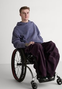 Lilla bomullshoodie med en virvel-logo, sammen med løse, mørkelilla bukser. Brukeren sitter i en rullestol med metall- og gummihjul.