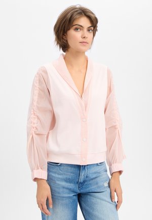 Jeune femme aux cheveux bruns courts portant un chemisier rose clair boutonné avec des manches froncées et un jean bleu sur un fond uni.