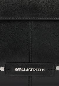 Sac à main en cuir noir avec une texture lisse, un design structuré, un matériel blanc bien visible et une étiquette de logo argentée.