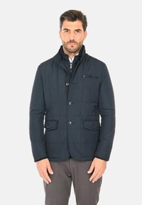 Giacca imbottita trapuntata blu navy con colletto alto, tasca sul petto con cerniera e due tasche frontali con patta. Tessuto liscio con design leggero.