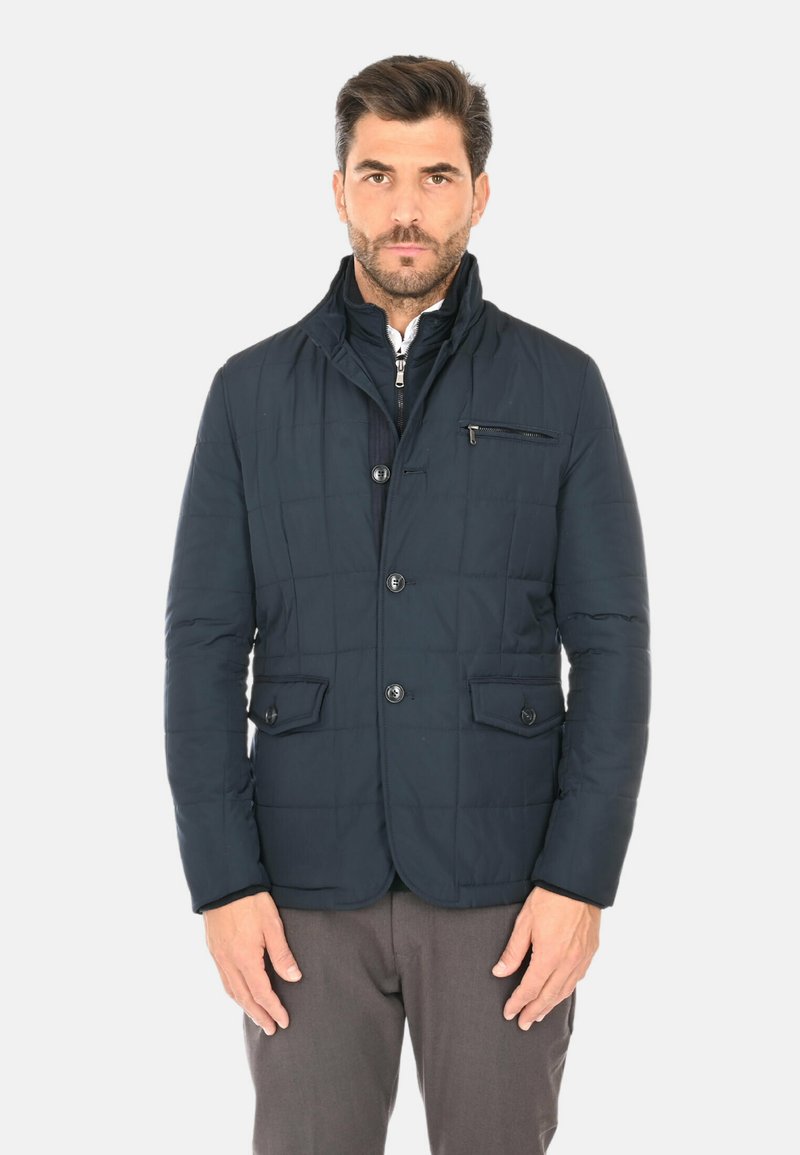 Giacca imbottita trapuntata blu navy con colletto alto, tasca sul petto con cerniera e due tasche frontali con patta. Tessuto liscio con design leggero.