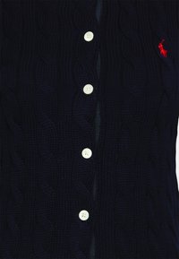 Marinblå stickad kofta med kabelmönster, vita knappar och en liten röd emblem på vänstra sidan. Mjuk textur och avslappnad passform.