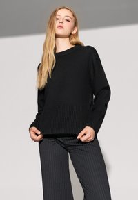 Czarny sweter z dzianiny z prążkowanym wykończeniem dołu i mankietów, o luźnym kroju i okrągłym dekolcie. Nosi się go z spodniami w pionowe paski.