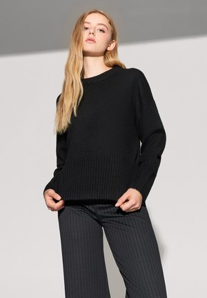 Maglione nero a maglia con orlo e polsini a coste, caratterizzato da una vestibilità comoda e un collo rotondo. Indossato con pantaloni a righe verticali.
