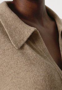 Beige stickad tröja med krage, som har en mjuk textur och ett ljust melerat mönster. Närbild av tyg och krags detaljer.