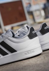 Baskets blanches Adidas avec des bandes noires et des patchs au talon, reposant sur une surface en béton avec un arrière-plan urbain flou.