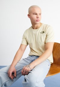 Camiseta de manga corta beige con un logo verde en el lado izquierdo del pecho, combinada con jeans de denim azul claro. Textura suave y corte casual.