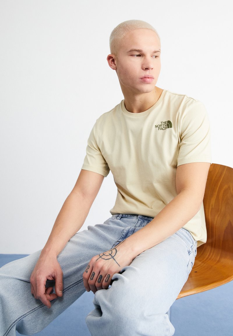 Camiseta de manga corta beige con un logo verde en el lado izquierdo del pecho, combinada con jeans de denim azul claro. Textura suave y corte casual.