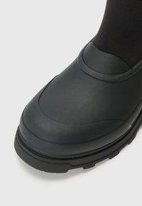Botte noire imperméable avec une finition lisse et mate, un bout rond et une semelle en caoutchouc texturé pour une meilleure traction. Conception de rebord supérieur ajustable.