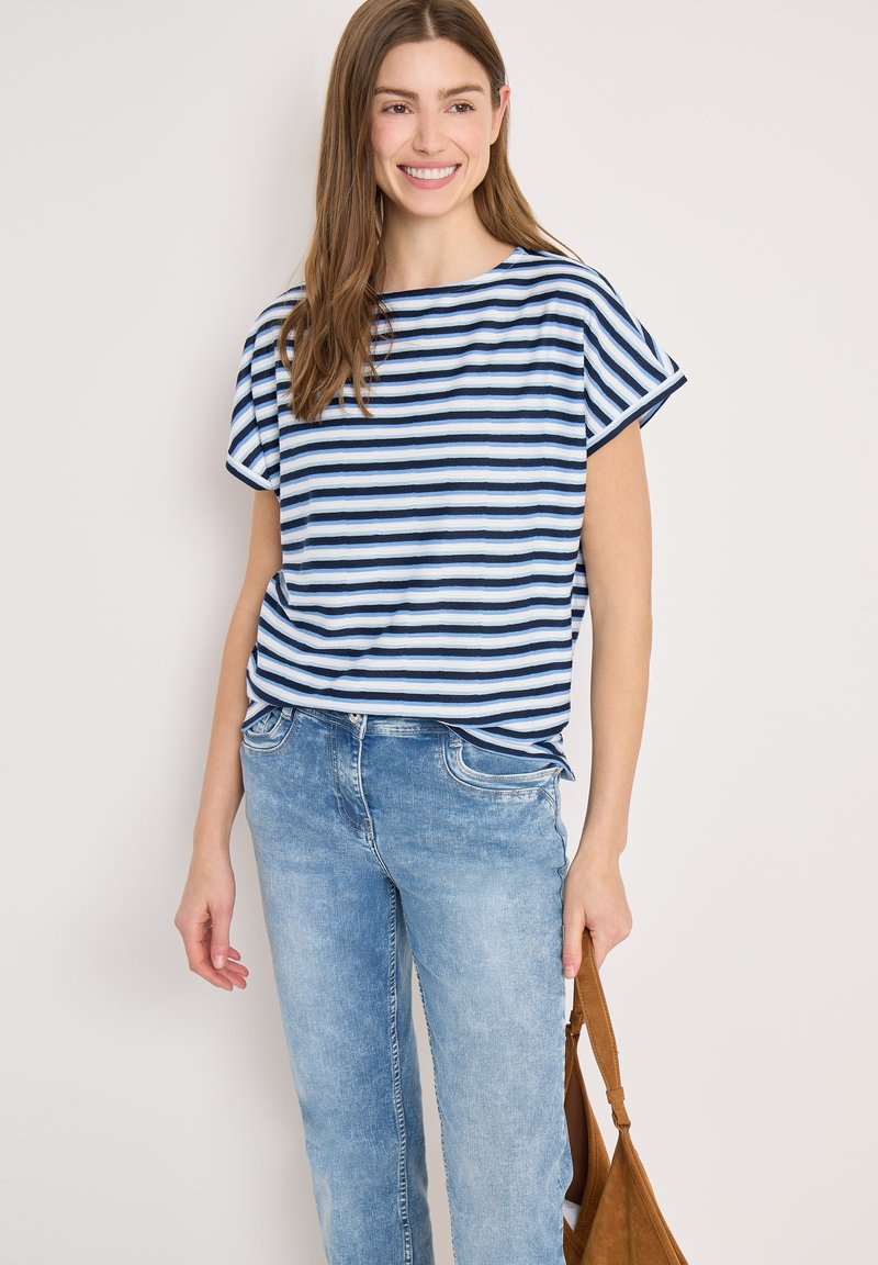 Femme souriante portant un haut à manches courtes rayé bleu et blanc et un jean bleu clair, tenant un sac à main en suède marron à ses côtés.