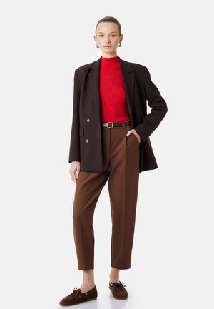 Woman stands with hand in pocket wearing red turtleneck, dark brown blazer, brown trousers, and brown moccasin shoes against white background.