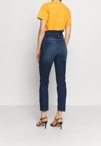Jeans de mezclilla azul oscuro de talle alto, con un corte ajustado, bolsillos traseros visibles, y complementados con una camiseta de manga corta naranja.