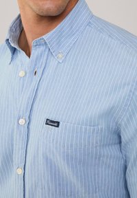 Camisa de un botón de color azul claro con rayas verticales blancas, un bolsillo en el pecho izquierdo y una pequeña etiqueta de marca en navy. Tejido de algodón suave, cuello clásico.