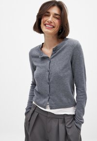 Gray knitted cardigan with a round neck, front buttons, and ribbed hem. Fabric appears soft and lightweight, worn over a white shirt.