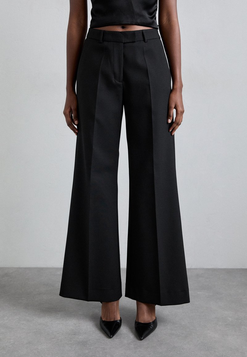 Sorte vide bukser med høj talje, struktureret stof og skarpe plisseringer. Stylet med spidse sorte hæle.