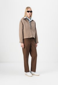 Brown cropped jacket with a light blue collar, paired with brown trousers and white sneakers. Model wearing black sunglasses.
