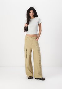 Pantaloni cargo bej, cu picioare largi, multiple buzunare și o croială lejeră, asortați cu o cămașă gri cu mâneci scurte. Textura netedă a materialului.