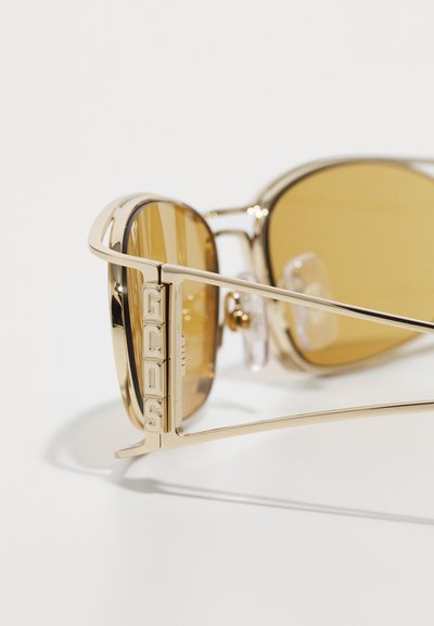 Gold-framed sunglasses with brown tinted lenses; features bold "GCDS" branding on the side and prominent temple design. Transparent nose pads included.