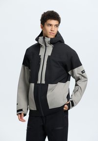 Giacca da uomo in nero e grigio chiaro. Presenta un cappuccio, tasche con zip e un mix di tessuti con finitura testurizzata. Inclusi polsini regolabili.