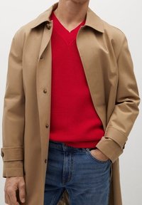 Beiger Trenchcoat mit Knopfdetails über einem roten V-Ausschnitt-Pullover, kombiniert mit blauen Jeans. Glatte Textur und klassische Designelemente.