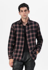 Camicia di flanella a quadri neri e rossi con colletto a camicia, tasca frontale e vestibilità rilassata, indossata sopra un top nero senza maniche. Pantaloni in pelle nera.