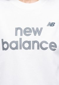 Λευκό πουκάμισο με το λογότυπο της New Balance και το κείμενο "new balance" σε περίγραμμα με ρίγες πάνω στο στήθος, καθώς και μικρό λογότυπο "NB" πάνω από το κείμενο.