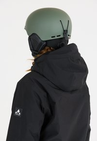 Zwarte waterdichte jas heeft een grote capuchon en een logo patch op de mouw. Een matte groene skibroek met verstelbare kinbanden wordt gedragen.