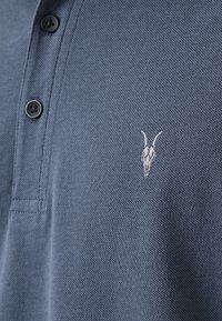 Mörkblå pikétröja med strukturerad väv, tre knappar och en vit broderad getskalle-logga på vänster bröst.