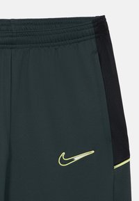 Pantalones cortos deportivos de color verde oscuro con paneles laterales de malla negra, cintura elástica y logo de Nike reflectante en color amarillo en el lado izquierdo.