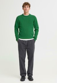 Zielony dziergany sweter z okrągłym dekoltem, noszony na białej koszulce; zestawiony z luźnymi ciemnoszarymi spodniami i czarnymi butami.