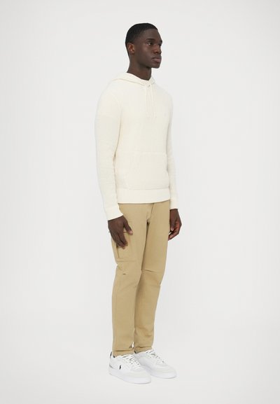 Krémová pletená mikina s kapucňou a predným vreckom, kombinovaná s béžovými cargo nohavicami a bielymi teniskami. Model stojí na jednoduchom pozadí.