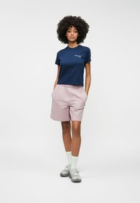 Marineblå bomulds-t-shirt med hvidt logo, parret med lyserøde ternede shorts. Modellen bærer grå sandaler og hvide ankelstrømper. Glat stof, afslappet pasform.